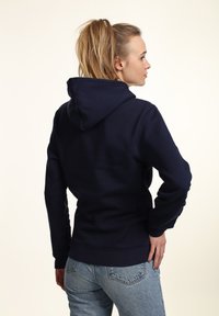 Hoodie blu navy con una texture liscia e cappuccio con cordini. Presenta maniche lunghe e una vestibilità aderente, abbinato a jeans azzurri chiari.