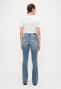 Felső részen bővülő, világoskék farmernadrág öt zsebbel, barna bőr folttal a deréknál, és elhalványult textúrával. Hátsó nézet.