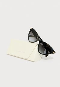 Melni kaķacu formas saulesbrilles ar gradienta lēcām, zelta akcenti uz tempļiem, atpūšas uz baltas, struktūrētas futrāles ar iepresētiem vārdiem "MICHAEL KORS."