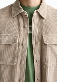 Camisa de pana beige con cuello con solapa, dos bolsillos frontales y cierre de botones, superpuesta sobre una camiseta verde con un pequeño logo.