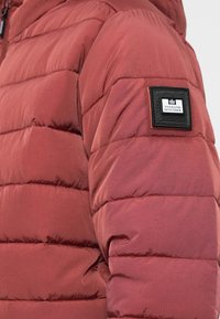 Rote Puffy-Jacke mit gestepptem Design, ausgestattet mit einem Logo-Patch auf dem linken Ärmel und einer glatten, reflektierenden Oberfläche.