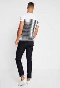 Camiseta de manga corta, blanca con rayas marinas, de diseño ajustado, llevada con jeans oscuros y zapatillas blancas. Vista trasera, textura suave.