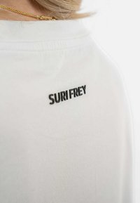 Bílý tričko z látky s krátkým rukávem, které na zádech u výstřihu zdobí malé černé vyšívané logo s nápisem "SURI FREY". Měkká textura.