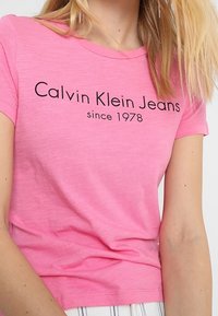 Růžové bavlněné tričko s krátkými rukávy, na kterém je vpředu vytištěno „Calvin Klein Jeans since 1978“ v černé barvě. Měkká textura a přiléhavý střih.