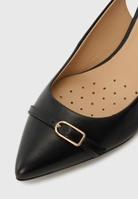 Gros plan d'une chaussure plate noire à bout pointu avec une petite bride à boucle dorée et une doublure intérieure beige perforée.