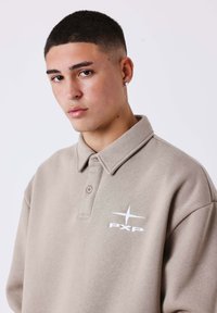 Polo shirt beige realizzata in tessuto morbido, con colletto e logo ricamato sul petto. Include una patta con bottoni e maniche corte.