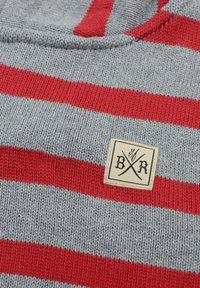 Grå stickad tröja med röda horisontella ränder. Har en fyrkantig logopatch märkt "B/R" i svart på en beige bakgrund. Mjuk textur.