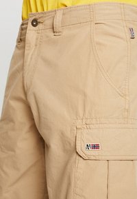 Pantaloni cargo beige in cotone con una texture liscia, dotati di una tasca laterale e di un piccolo logo ricamato.