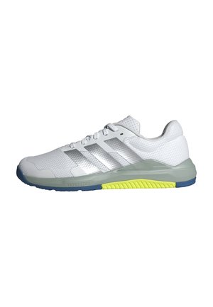 Zapatilla deportiva blanca con rayas plateadas, entresuela gris, detalle de dibujo amarillo, suela exterior azul y cordones blancos sobre un fondo blanco liso.