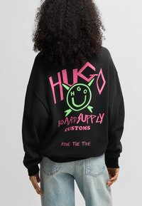 Felpa nera oversized con testo grafico audace in rosa e verde: "HUGO BOARD SUPPLY CUSTOMS" e un design con faccina sorridente.