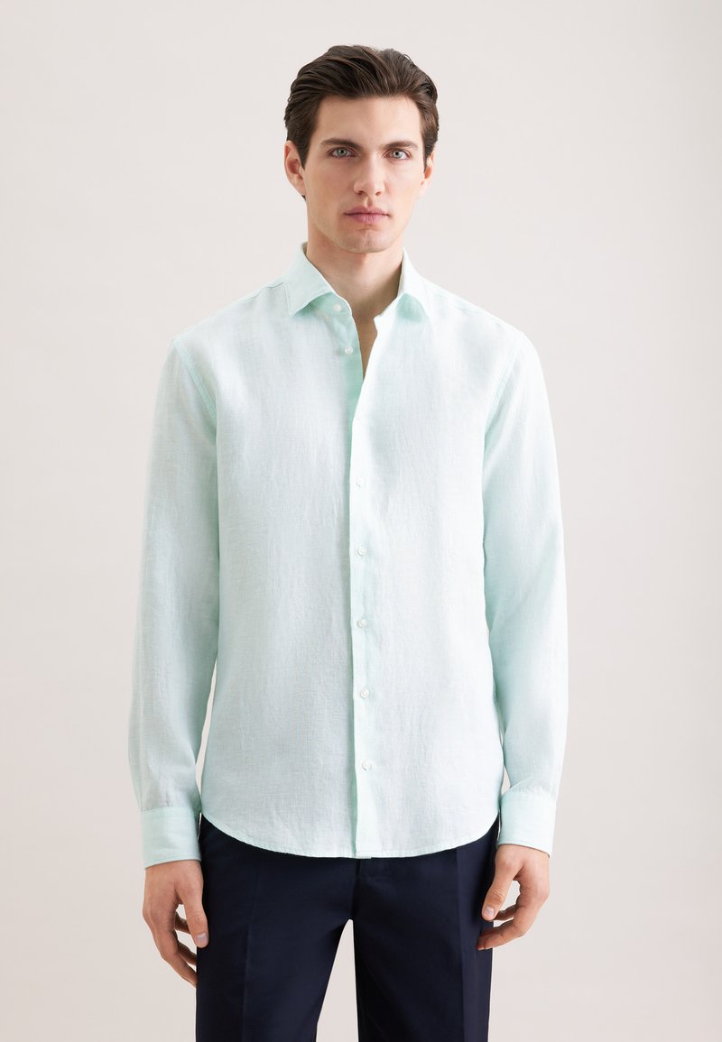 Jeune homme regardant vers l'avant, portant une chemise à manches longues bleu clair boutonnée et un pantalon foncé, sur un fond uni.