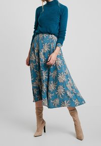 Kvinde iført teal turtleneck sweater, blå paisley midi nederdel og beige suede højhælede støvler, stående mod en ensfarvet baggrund.