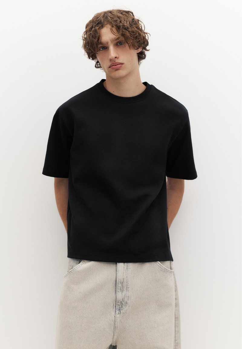 Jeune homme aux cheveux bouclés portant un t-shirt noir ample et un jean clair, debout devant un fond blanc uni.