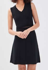 Robe noire sans manches avec un col en V festonné, une taille cintrée, une jupe évasée et des détails subtils de coutures verticales. Matière en maille douce.