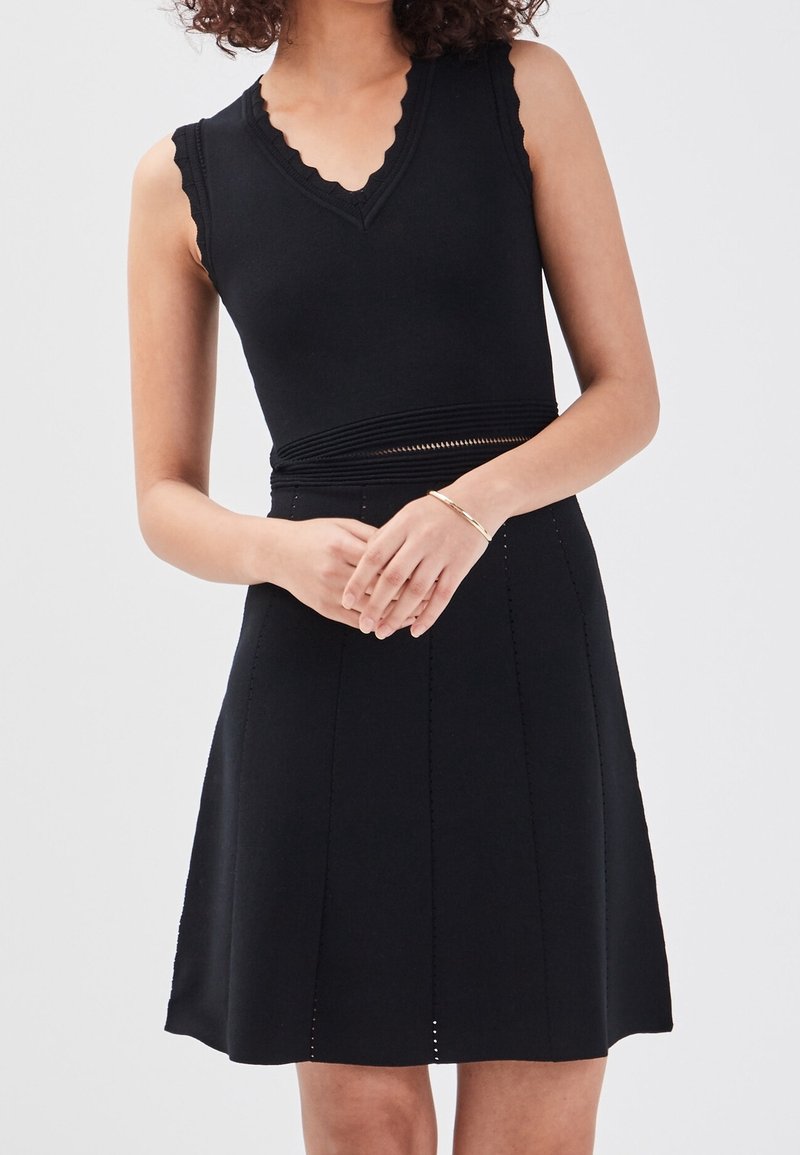 Robe noire sans manches avec un col en V festonné, une taille cintrée, une jupe évasée et des détails subtils de coutures verticales. Matière en maille douce.