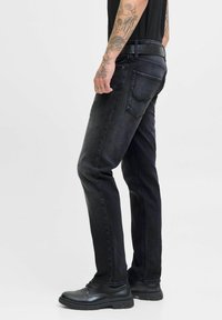 Hombre que lleva puesto unos pantalones negros ajustados, un cinturón negro, zapatos de cuero negros y una camisa negra, con el brazo izquierdo tatuado visible.