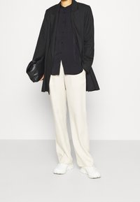 Sort frakke, mørk skjorte, cremefarvede vide bukser og hvide sneakers. Modellen holder en sort taske og står foran en ensfarvet baggrund.