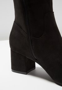 Bottine en daim noir avec talon carré, fermeture éclair latérale et texture lisse. Le design présente une discrète couture courbée en accent.