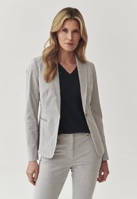 Gestreifter Blazer aus leichtem Stoff mit tailliertem Schnitt. Kombiniert mit einem dunklen V-Ausschnitt-Oberteil. Vertikale Streifen sowohl auf dem Blazer als auch auf der passenden Hose.
