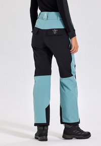 Black and teal ski pants featuring a high waist, zippered side pockets, and reinforced lower legs, paired with black hiking boots.