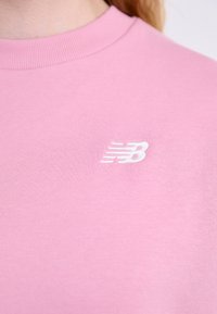 Sweatshirt rose clair avec un col rond, arborant un logo New Balance brodé blanc sur la poitrine. Matériau en coton doux, texture lisse.