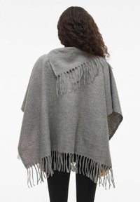 Sieviete ar tumšiem cirtainiem matiem, valkājot pelēku vilnas šalli ar franjām pār melniem biksēm, attēlota no mugurpuses uz balta fona.