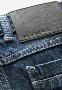 Jeans de mezclilla oscura con un parche de cuero negro texturizado en la parte trasera, con detalles de costura y un pequeño bolsillo. Se pueden ver costuras de color dorado claro.