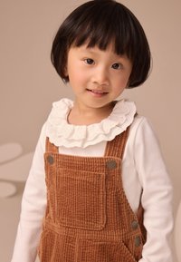 Salopette in velluto a coste marrone con tasca frontale, bottoni metallici e camicia bianca con colletto arricciato. Capelli corti neri. Sfondo neutro.