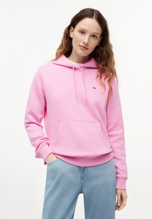 Ung kvinde med langt hår iført en lyserød hættetrøje med frontlomme og lyseblå jeans, stående mod en ensfarvet baggrund.