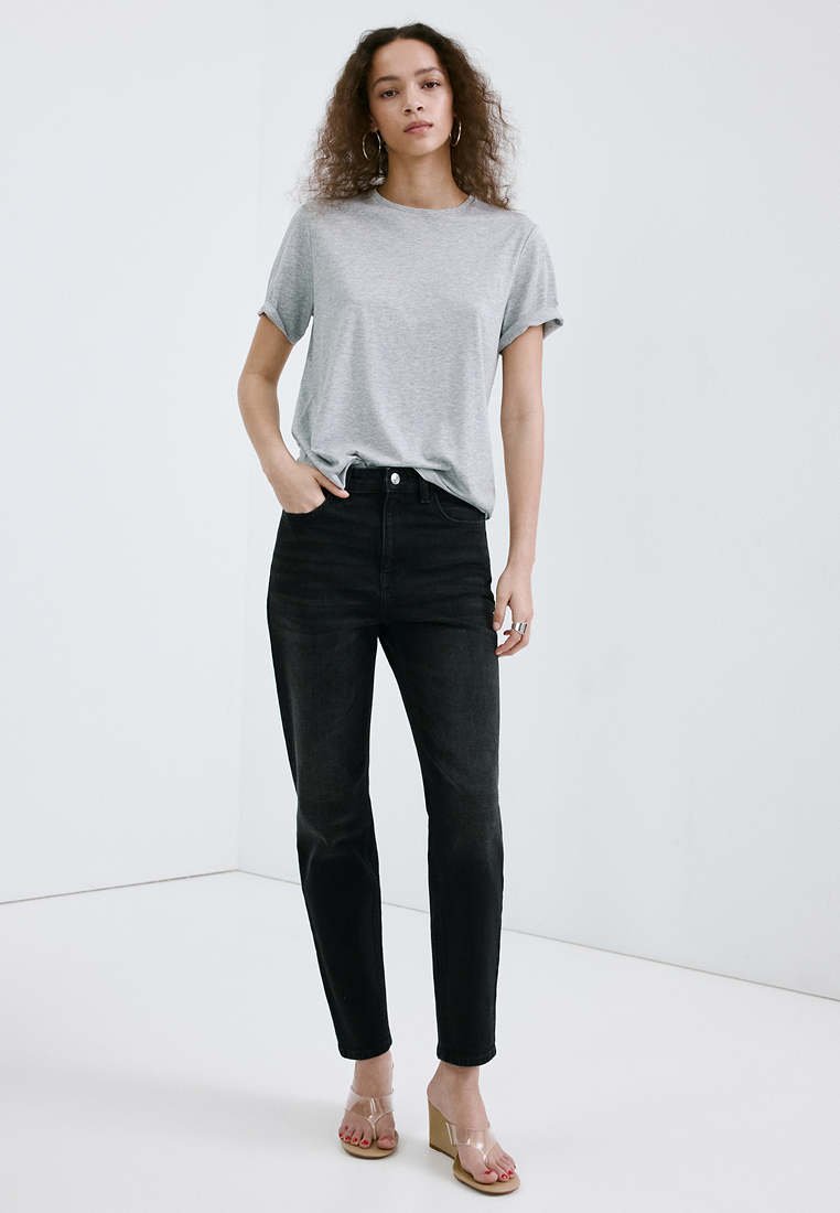 Femme aux cheveux bouclés, debout, portant un t-shirt gris clair, un jean noir taille haute, des sandales compensées transparentes et des boucles d'oreilles créoles.