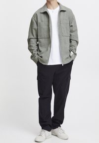 Grijze jas met rits en twee zakken aan de voorkant, gedragen over een wit T-shirt, gecombineerd met zwarte cargobroek en witte sneakers.