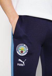 Marineblaue Jogginghosen mit hellblauen Seitenflächen, versehen mit einem Manchester City FC Logo und einem Puma-Logo. Hergestellt aus strukturiertem Stoff mit Taschen.