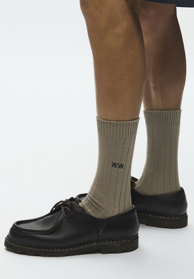 Jambes portant des chaussettes beiges côtelées avec le logo noir "WAW" et des chaussures en cuir noires à lacets sur un fond clair uni.