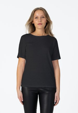 Femme aux longs cheveux blonds portant un t-shirt noir uni à manches courtes et un pantalon en cuir noir, debout devant un fond blanc.