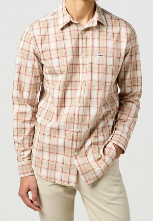 Hombre con camisa de manga larga a cuadros beige y roja con bolsillo en el pecho y pantalones beige, con la mano en el bolsillo.