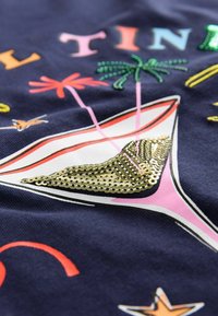 Marineblåt stof med farverigt, broderet cocktailglasdesign, der har guldpalietdetaljer, grønne palmetræer og stjerneakcenter.
