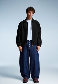 Veste bomber noire avec des poignets côtelés, t-shirt blanc en dessous, et pantalon en denim bleu ample. Des baskets bordeaux complètent la tenue.