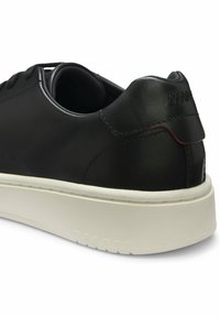 Basket en cuir noir avec semelle blanche épaisse, design bas, lacets noirs et détail subtil du logo au talon, vue de l'arrière sur le côté.