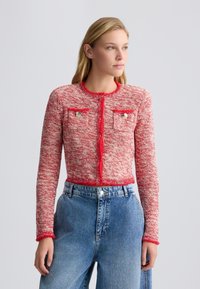 Cardigan in tweed rosso con maniche lunghe, dotato di due tasche frontali e bottoni argentati. Rifinito con dettagli pelosi rossi per un tocco di texture. Indossato con denim.