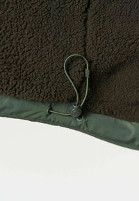 Gros plan de l'ourlet d'une veste polaire vert foncé avec un cordon élastique réglable et un bouton en plastique.