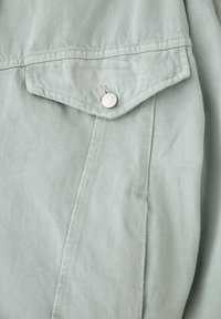 Vêtement vert clair en tissu texturé, avec un col pointu et une finition avec bouton-pression argenté sur la poche poitrine.