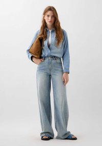Lichtblauw denim shirt met knopen, hoog-idiots lichtblauwe jeans en zwart-blauwe sandalen. Bruine tas onder de arm, neutrale achtergrond.