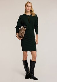 Abito in maglia verde fitted con maniche lunghe, abbinato a stivali neri lucidi al ginocchio e una borsa marrone. Una collana con pendente dorato completa l'outfit.