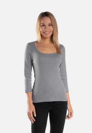 Uśmiechnięta kobieta o prostych blond włosach, ubrana w szary, dopasowany top z długim rękawem i czarne spodnie, stojąca na jednolitym tle.