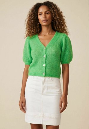 Femme aux cheveux bouclés portant des boucles d’oreilles en anneau dorées, un pull boutonné manches courtes vert vif et une jupe blanche longueur genou.