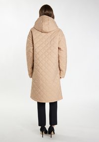 Beige gewatteerde lange jas met een capuchon, voorzien van ruitvormige stiksels en een comfortabele pasvorm. Gepaard met zwarte broek en hoge hakken.