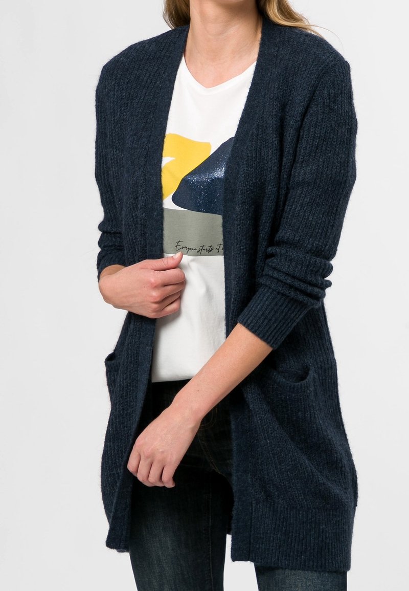 Cardigan en maille bleu marine avec devant ouvert, manches longues et deux poches avant, porté sur un t-shirt graphique blanc. Tissu texturé.