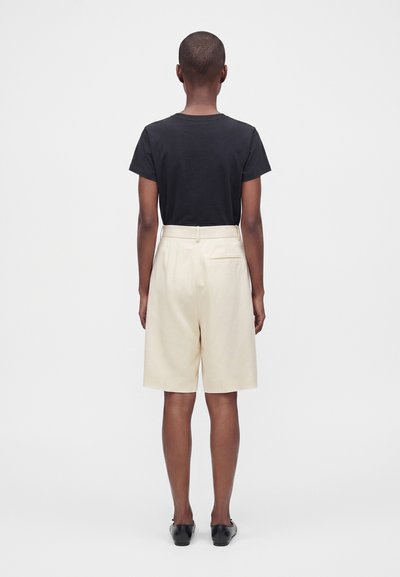 Personne debout de dos, portant un t-shirt noir, un short beige aux genoux et des chaussures noires sans lacets sur un fond uni.