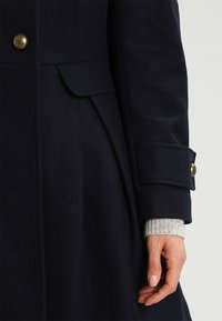 Abrigo de lana azul con herrajes de botones dorados, una silueta ajustada y puños de punto gris texturizados visibles en la muñeca.