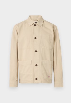 Lichtbeige werkjasje, katoenen stof, knoopsluiting aan de voorkant, puntige kraag, twee borstzakken, twee lagere zakken, bruine knopen, gestikte details.